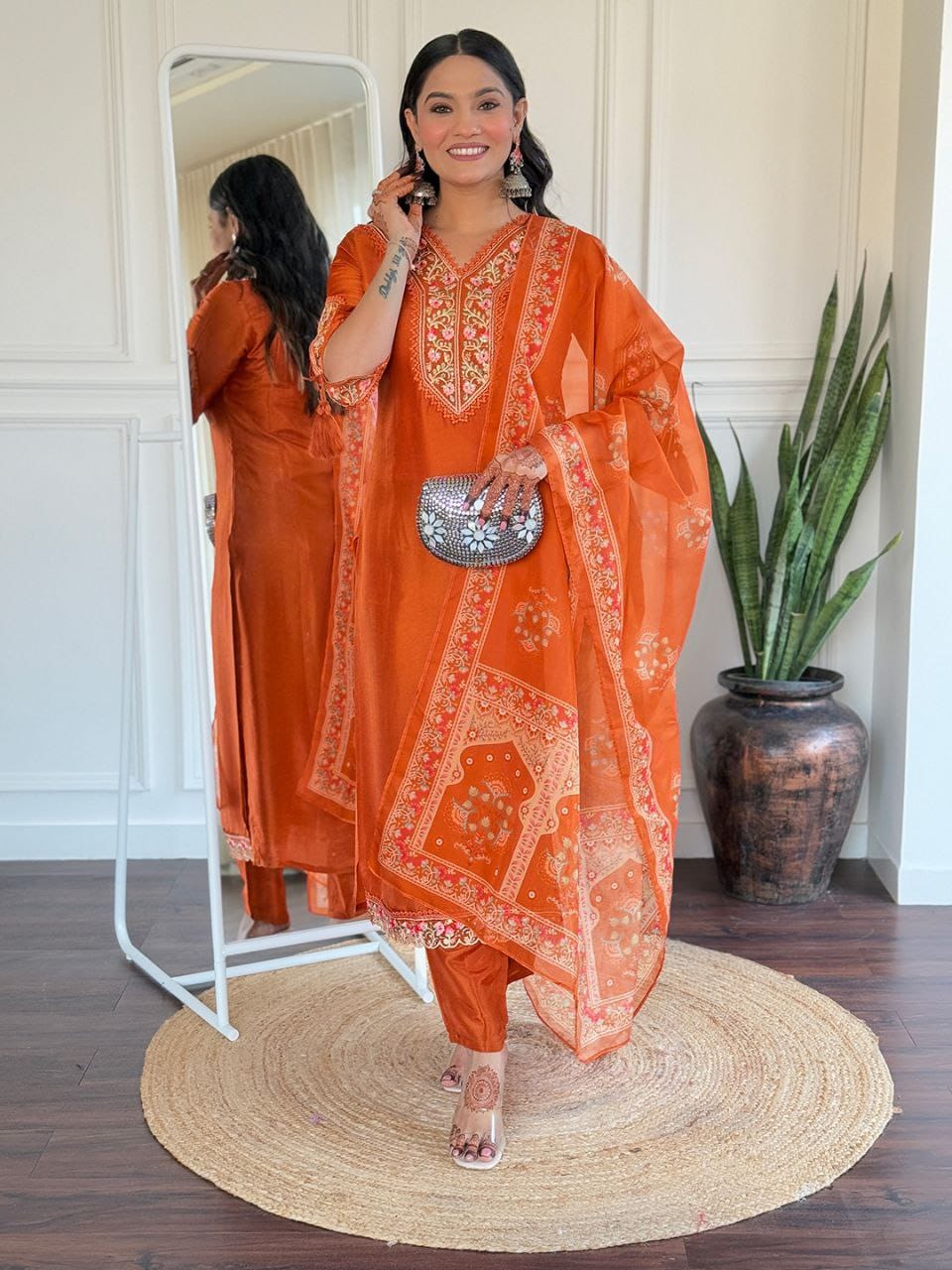 Woman in an orange traditional outfit standing in a room with a mirror and plant.