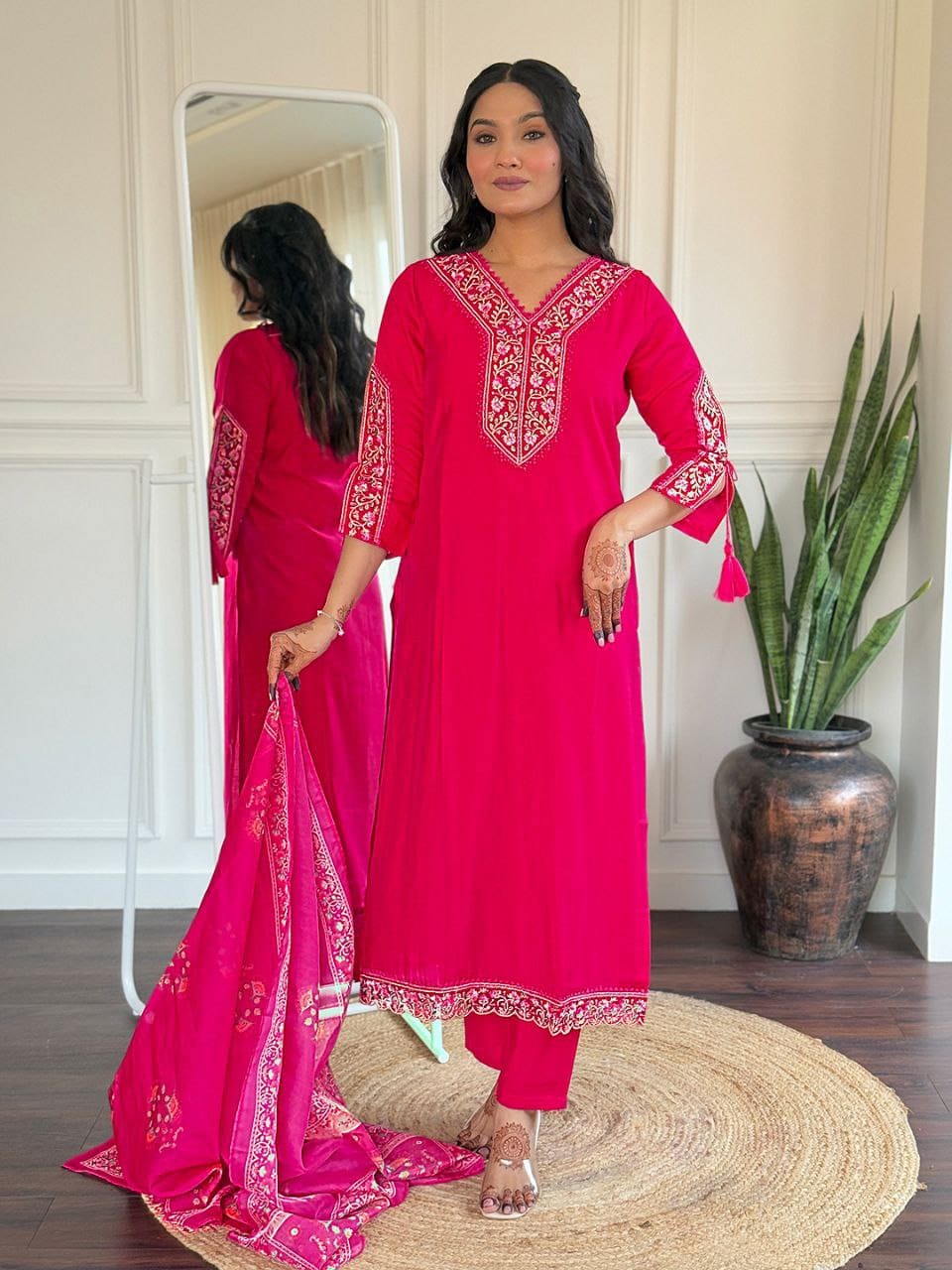 Woman in a bright pink traditional outfit with intricate designs, standing in a room with a mirror and plant.