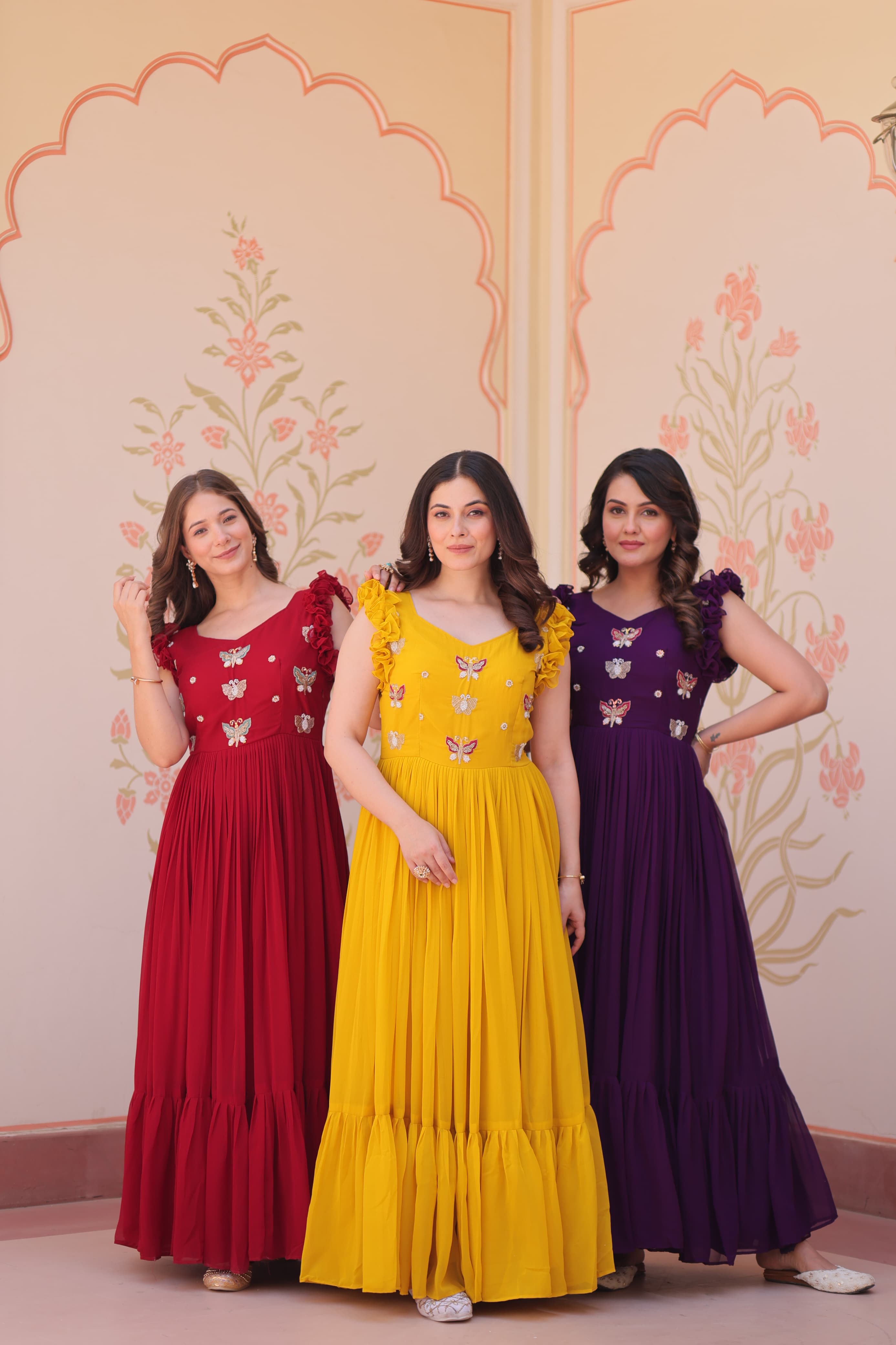 Three women in colorful long dresses standing against a decorative wall.