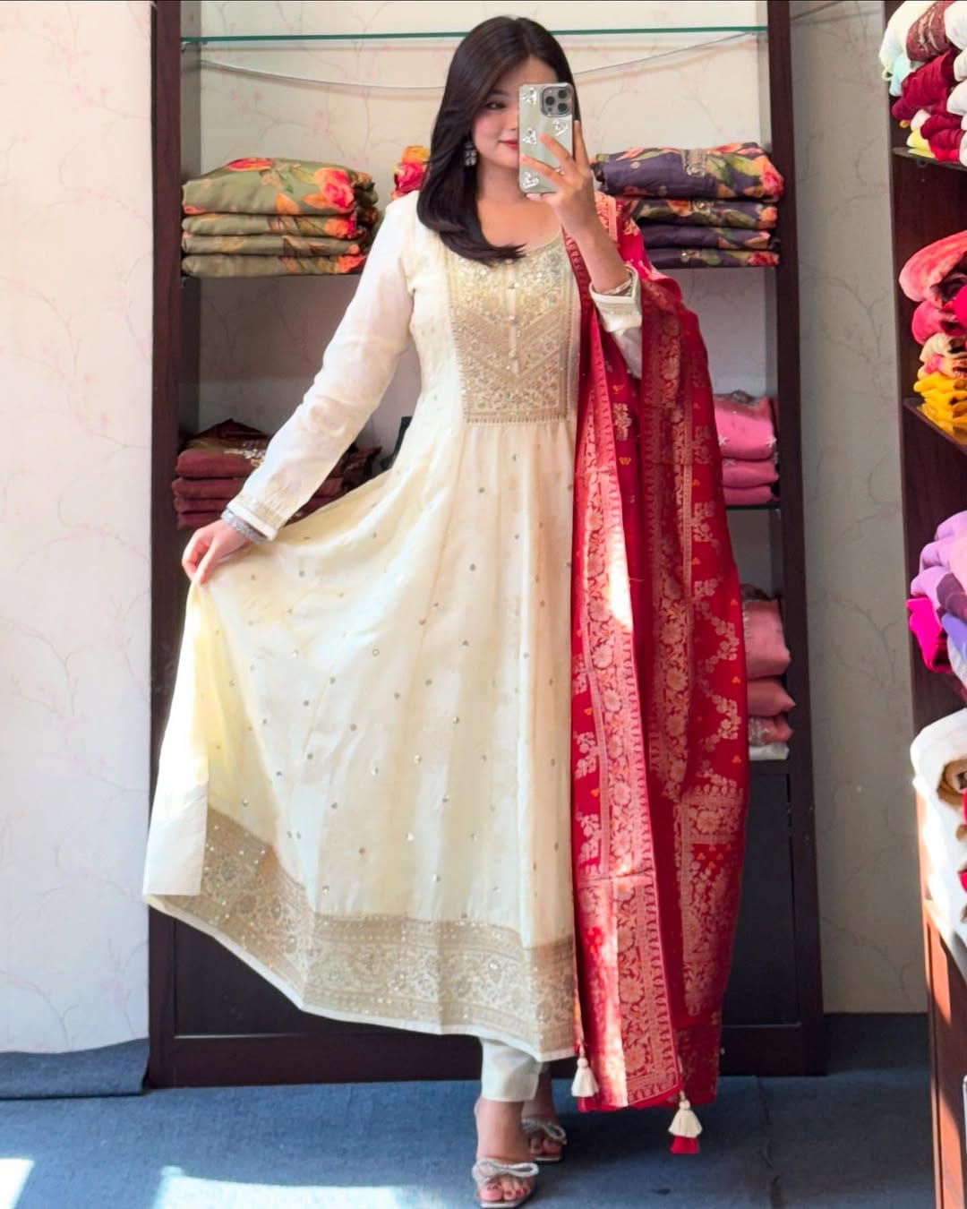 Woman in a white traditional outfit with a red dupatta taking a mirror selfie in a store.